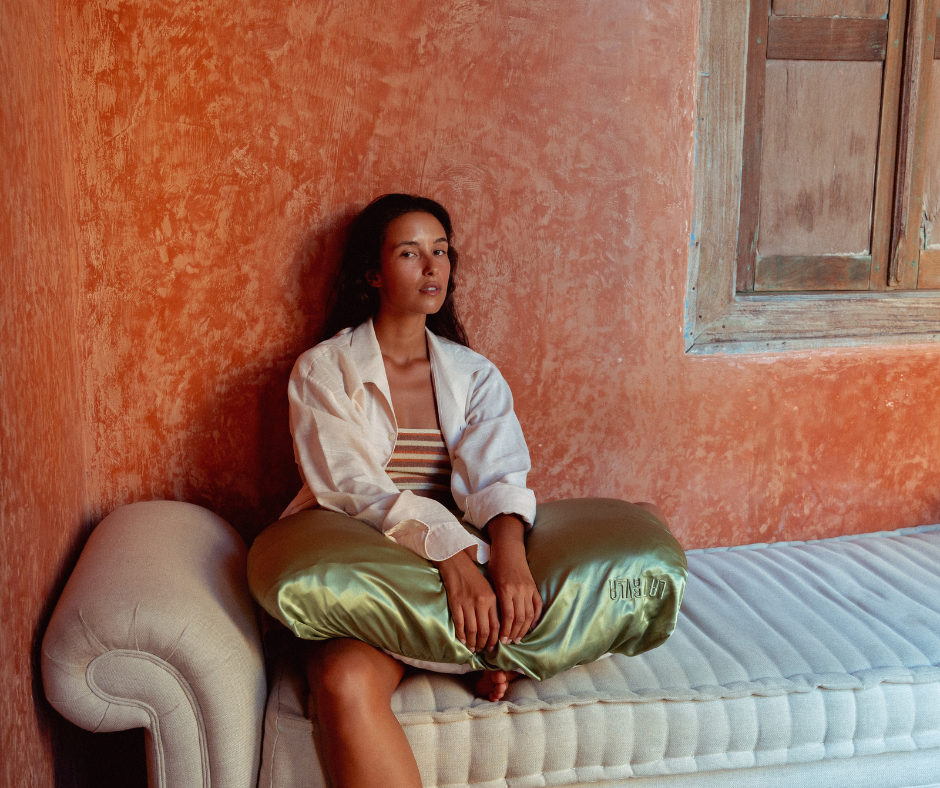 Woman sitting on a bed with a textured orange wall and wooden window frame holding the TUNDRA PillowWrap by LATRAVLA.
