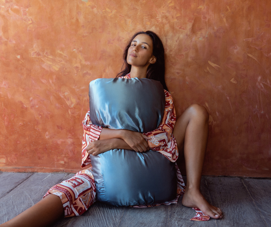 Woman sitting on the floor holding a large steel blue mulberry silk LATRAVLA PillowWrap against an orange wall.